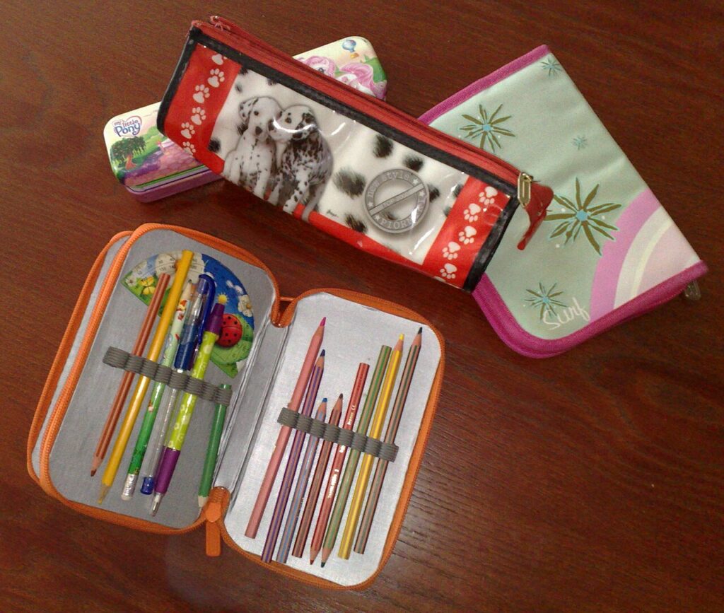 Several pencil cases on a wooden table, including an open orange-trimmed case with pencils and pens held in elastic loops.