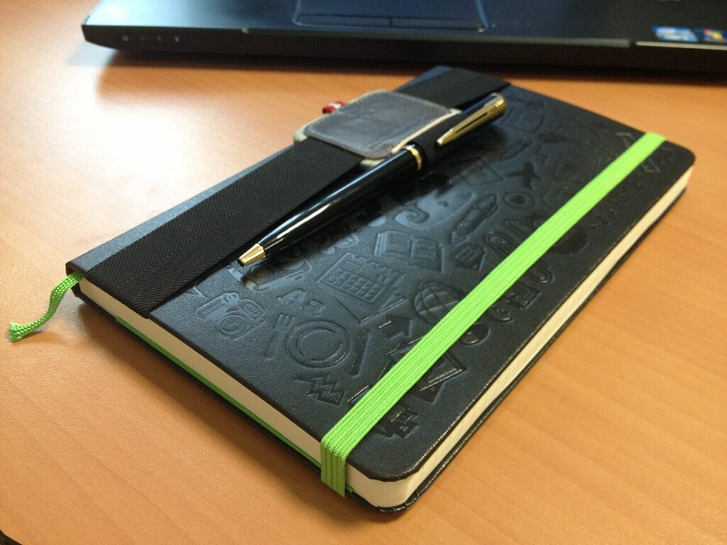 Black notebook with a bright green elastic closure and a black-and-gold pen resting across a wide black strap.