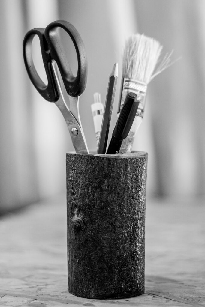 Black-and-white photo of a cylindrical holder containing scissors, a pencil, pens, and a paintbrush.