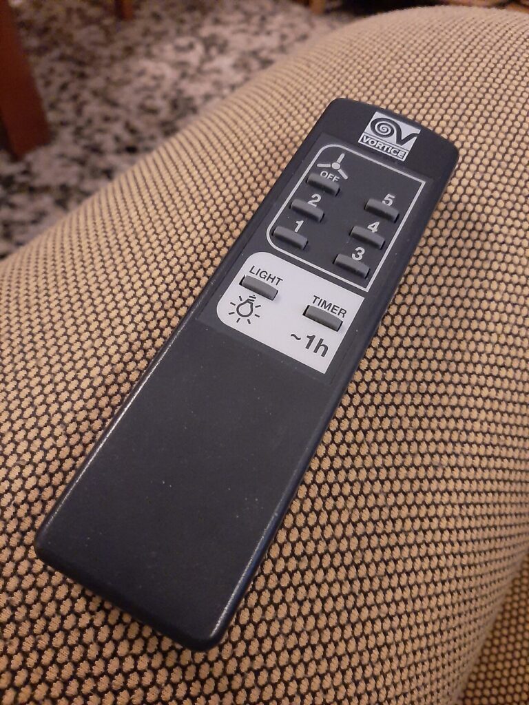 Black Vortice fan remote with numbered speed buttons, light and timer controls, resting on patterned upholstery.